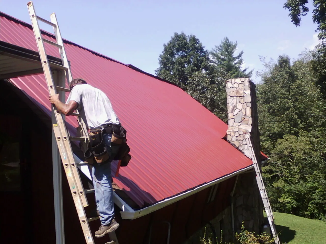 Roofer on a ladder beside a red metal roof and stone chimney — Soffit & Fascia Repair by Roofing Brothers in Elk Falls
